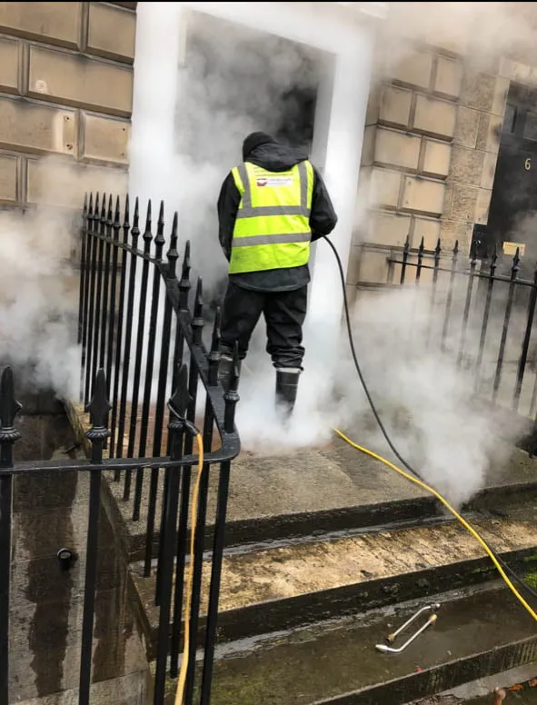 Worker in high-visibility clothing steam-cleaning stonework and iron railings
