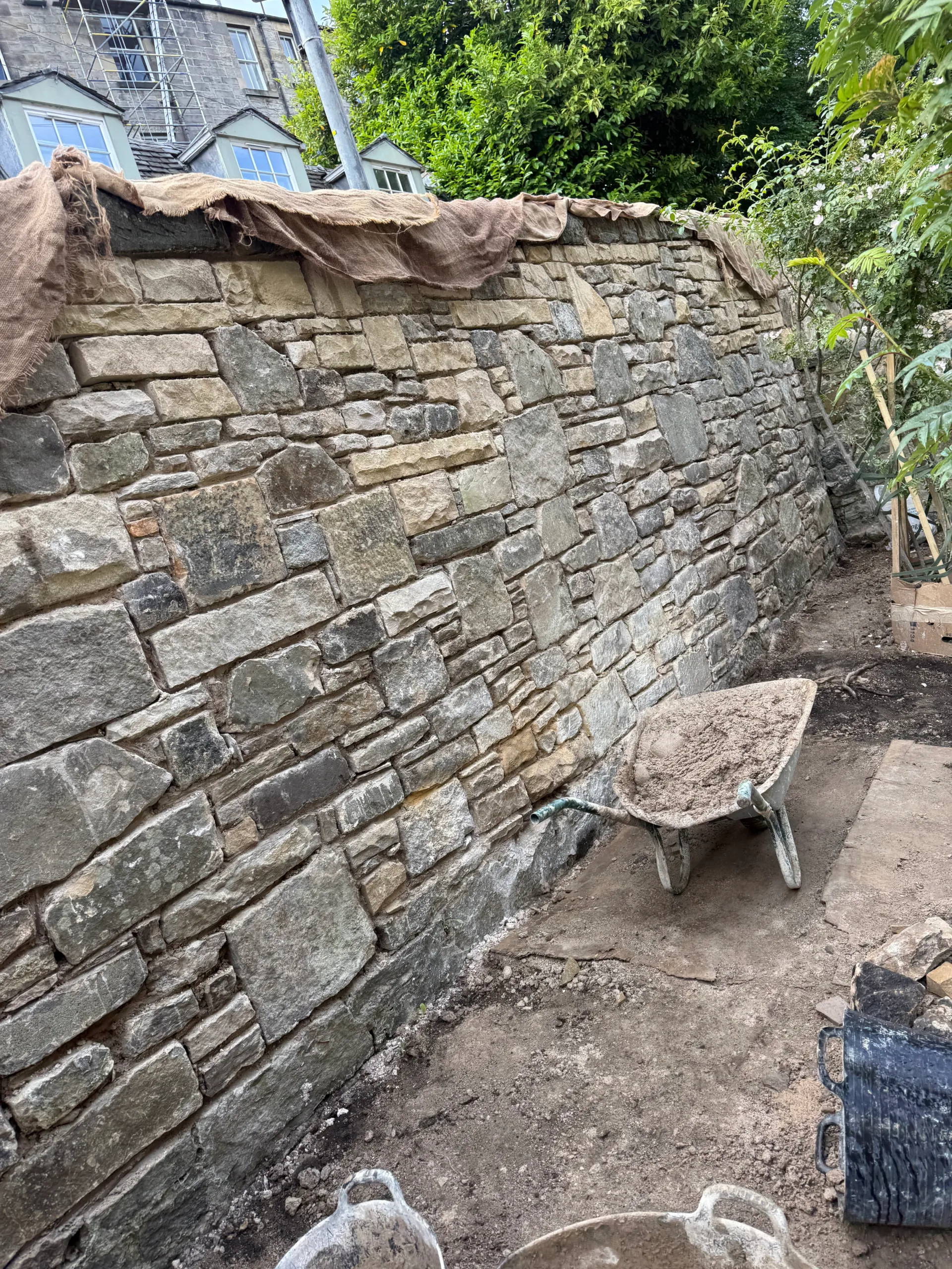 Stone garden wall part-way through rebuilding, with a wheelbarrow and covering sheet