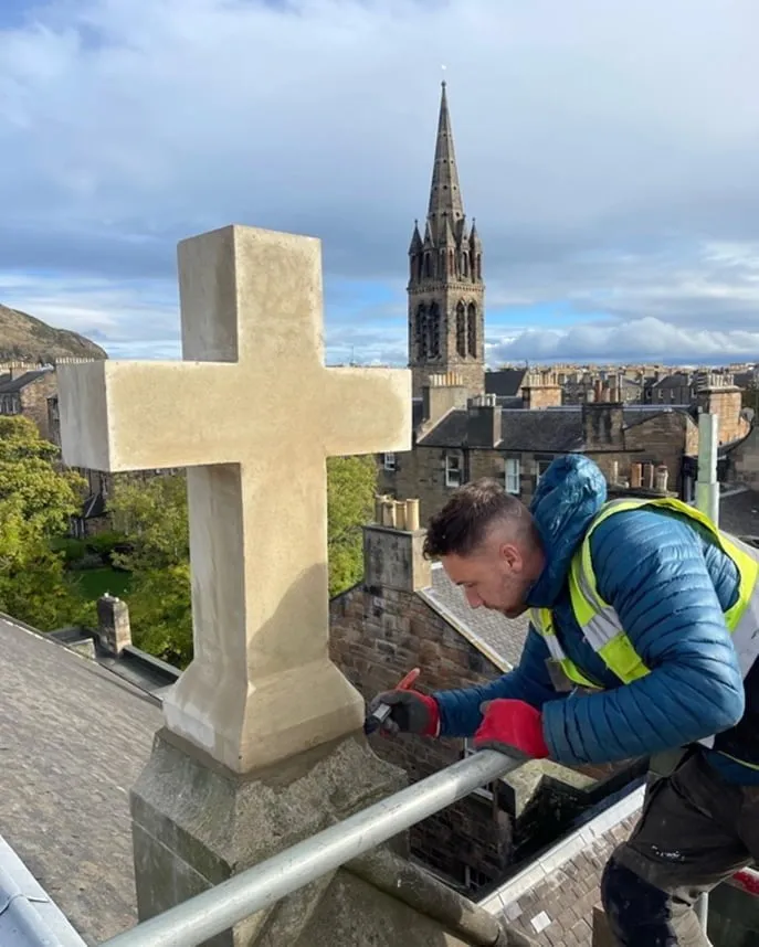 Stonemason repairing a carved stone cross at roof level, with a church spire behind