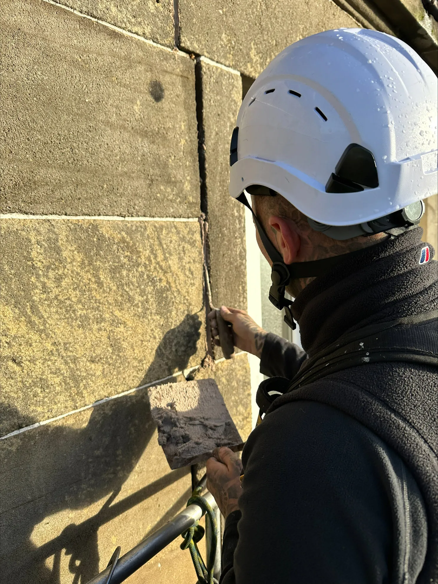 Stonemason in a helmet raking out a mortar joint in an ashlar wall